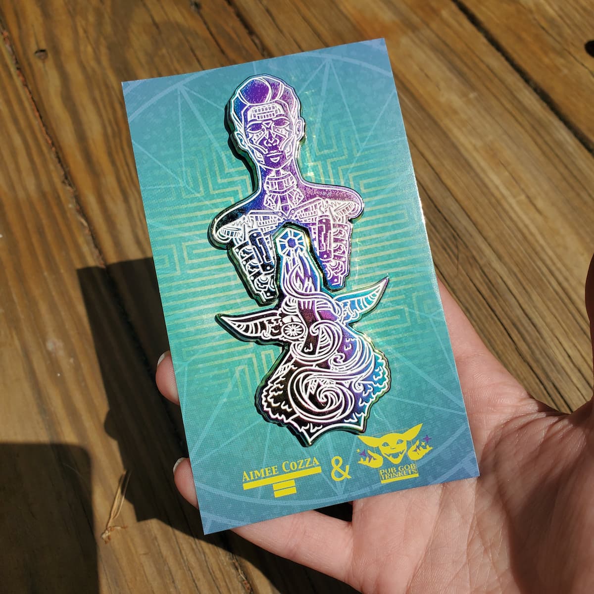 A hand holding a blue, green, and yellow card with two anodized pins on it of a robot torso, and a goblin holding a gemstone up into the heart of it.