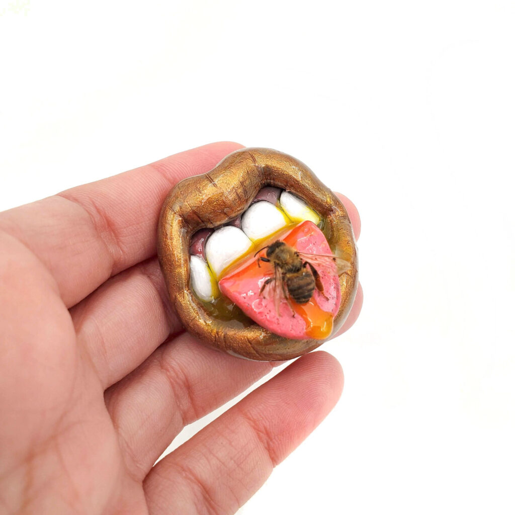 A polymer mouth with copper/gold lips, honey dripping out of the mouth and tongue, and a real honeybee on the tongue. It's held in a human hand.