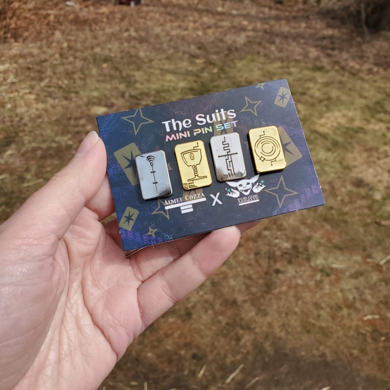 A white woman's hand holding a small backing card with 4 card-shaped pins on them, in silver and gold. It reads "The Suits Mini Pin Set"